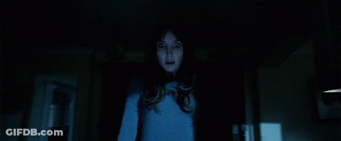 A Woman In A Blue Shirt Is Standing In A Dark Room Looking At The Camera GIF