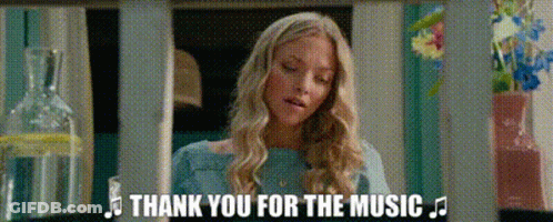 A Woman In A Blue Shirt Is Standing In Front Of A Bottle Of Water And Flowers And Says Thank You For The Music . GIF