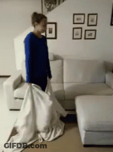 A Woman In A Blue Shirt Is Standing In Front Of A White Couch Holding A White Sheet GIF