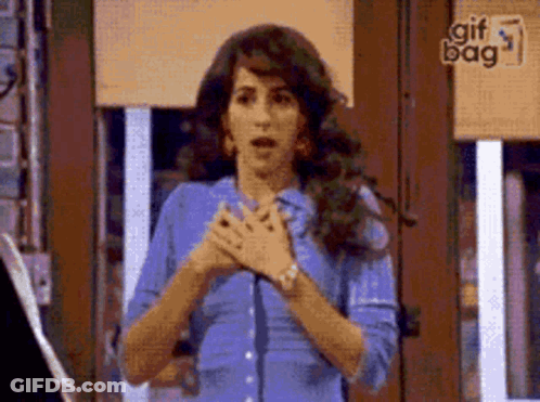 A Woman In A Blue Shirt Is Standing In Front Of A Window With Her Hands Folded In Front Of Her Chest . GIF