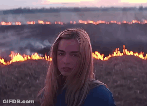 A Woman In A Blue Shirt Stands In Front Of A Field Of Fire GIF