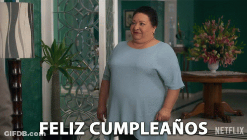 A Woman In A Blue Shirt Stands In Front Of A Sign That Says " Feliz Cumpleanos " GIF