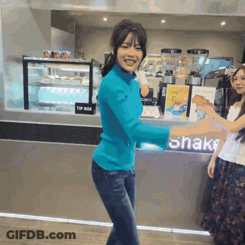 A Woman In A Blue Shirt Stands In Front Of A Sign That Says Shake GIF