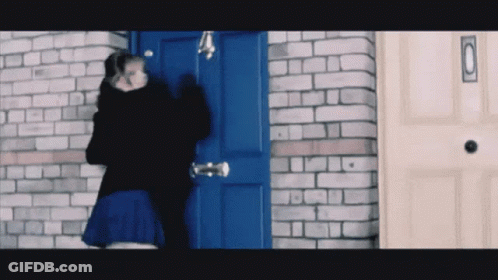 A Woman In A Blue Skirt Stands In Front Of A Blue Door With A Brick Wall Behind Her GIF