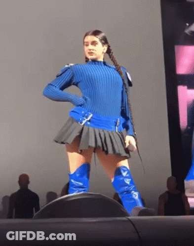 A Woman In A Blue Sweater And Blue Boots Stands On A Stage Holding A Microphone GIF