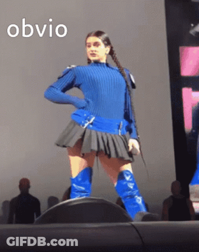 A Woman In A Blue Sweater And Blue Boots Stands On A Stage With The Word Obvio Written Above Her GIF