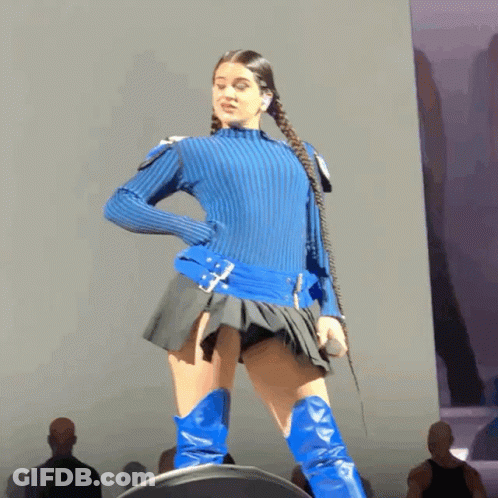 A Woman In A Blue Sweater And Blue Boots Stands On Stage GIF