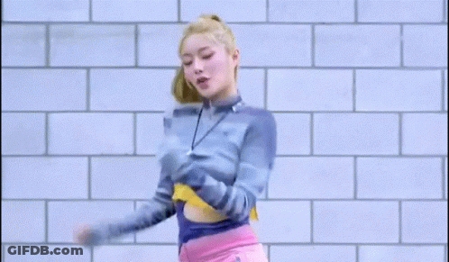A Woman In A Blue Sweater And Pink Shorts Is Dancing In Front Of A Brick Wall . GIF