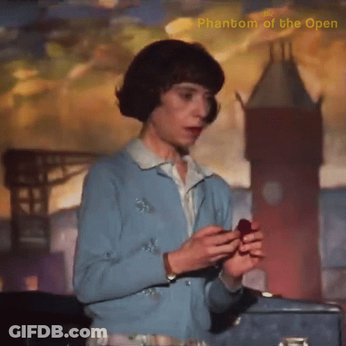 A Woman In A Blue Sweater Is Holding A Red Object In Front Of A Phantom Of The Open Painting GIF