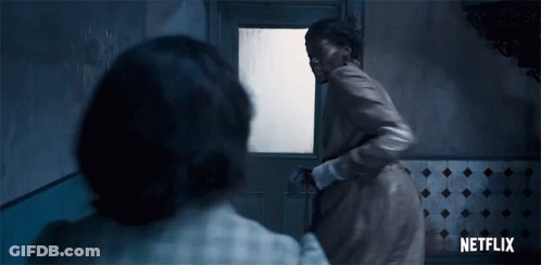 A Woman In A Brown Coat Is Standing In Front Of A Door In A Room . GIF