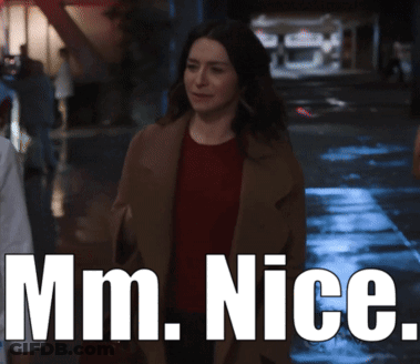 A Woman In A Brown Coat Is Walking Down A Street With The Words Mm Nice Behind Her GIF