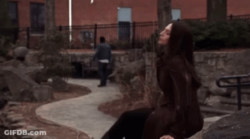 A Woman In A Brown Coat Sits On A Rock While A Man Walks By GIF