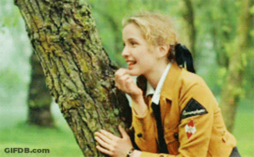 A Woman In A Brown Jacket Is Leaning Against A Tree Trunk And Smiling GIF