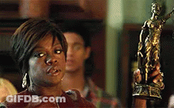 A Woman In A Brown Jacket Is Standing In Front Of A Statue Of A Woman . GIF