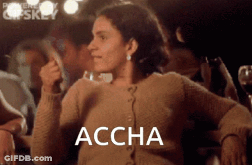A Woman In A Brown Sweater Is Sitting In Front Of A Group Of People And The Word Accha Is On The Screen GIF