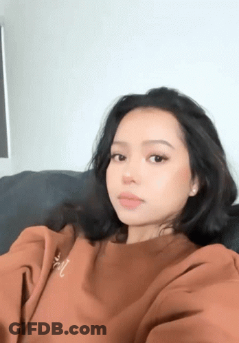 A Woman In A Brown Sweatshirt Is Sitting On A Couch Taking A Picture Of Herself . GIF