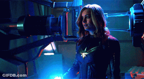 A Woman In A Captain Marvel Costume Is Standing In Front Of A Blue Light GIF