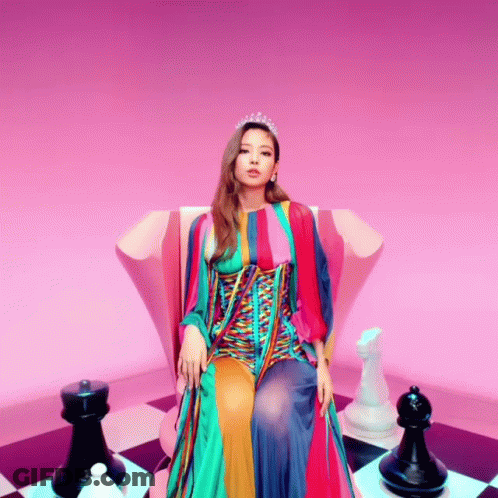 A Woman In A Colorful Dress Is Sitting On A Chair With Chess Pieces Around Her GIF