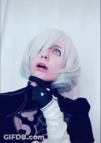 A Woman In A Cosplay Costume With White Hair And Black Gloves Is Standing In Front Of A White Wall . GIF