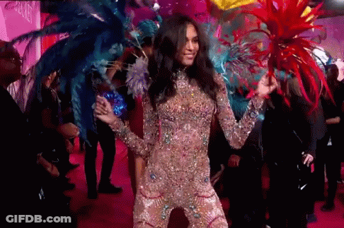 A Woman In A Costume With Feathers On Her Head Is Dancing On A Pink Carpet . GIF