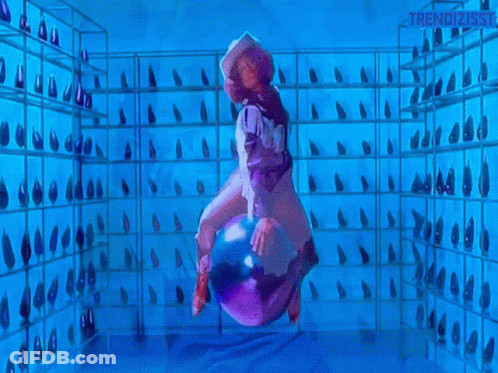 A Woman In A Cowboy Hat Is Riding A Purple Object In A Room With A Lot Of Shoes On Shelves . GIF