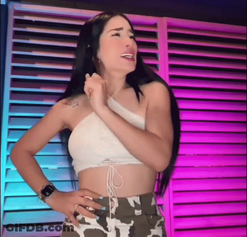 A Woman In A Crop Top And Camo Shorts Is Standing In Front Of A Purple Wall . GIF