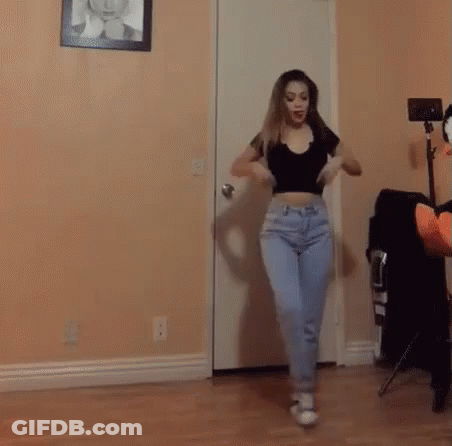 A Woman In A Crop Top And Jeans Is Dancing In A Room . GIF