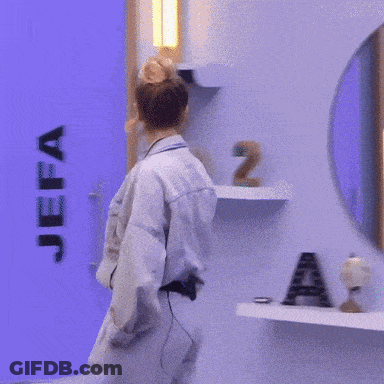A Woman In A Denim Jacket And Pants Stands In Front Of A Blue Wall GIF
