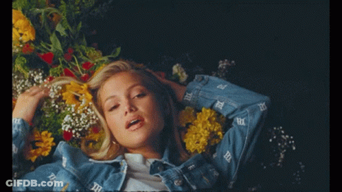 A Woman In A Denim Jacket Is Laying In A Bed With Flowers GIF