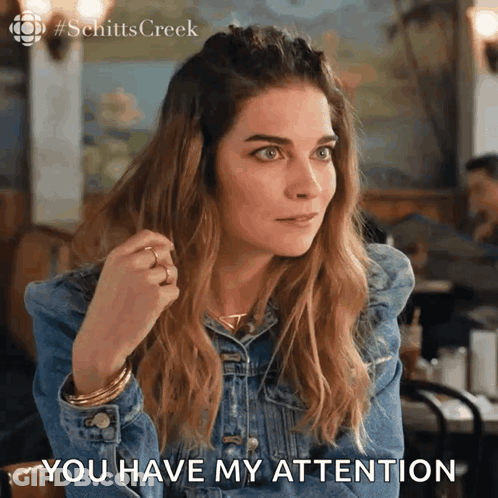 A Woman In A Denim Jacket Is Sitting At A Table And Says " You Have My Attention " GIF