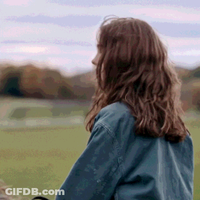 A Woman In A Denim Jacket Is Standing In A Field With Her Back To The Camera GIF