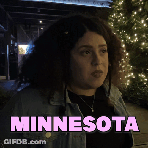 A Woman In A Denim Jacket Is Standing In Front Of A Sign That Says Minnesota GIF