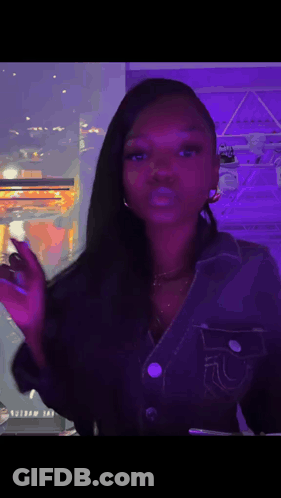 A Woman In A Denim Jacket Is Standing In Front Of A Window With A Purple Light Behind Her . GIF