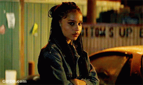 A Woman In A Denim Jacket Is Standing Next To A Car With Her Arms Crossed And Looking At The Camera . GIF