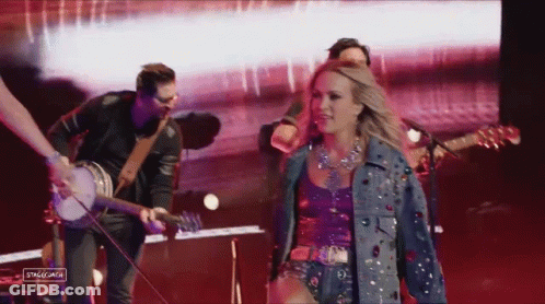 A Woman In A Denim Jacket Is Walking On Stage With A Band GIF