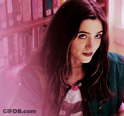 A Woman In A Denim Jacket Stands In Front Of A Bookshelf And Looks At The Camera GIF