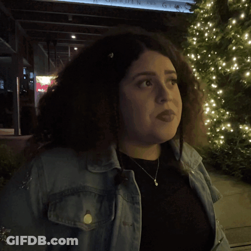 A Woman In A Denim Jacket Stands In Front Of A Sign That Says What GIF