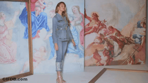 A Woman In A Denim Jacket Stands In Front Of A Wall Of Paintings GIF