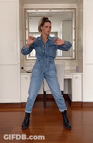 A Woman In A Denim Jumpsuit And Black Boots Is Dancing In Front Of A Mirror GIF