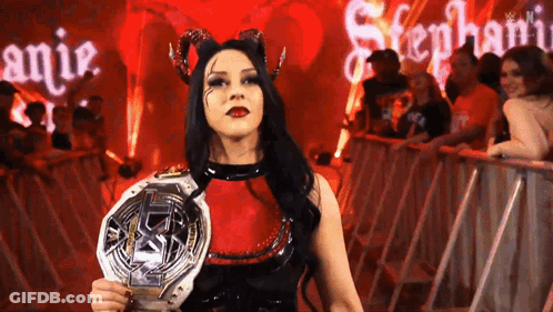 A Woman In A Devil Costume Is Holding A Championship Belt In Front Of A Crowd . GIF