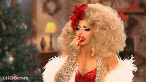 A Woman In A Drag Queen Costume Is Sitting In Front Of A Christmas Tree And Eating Something . GIF