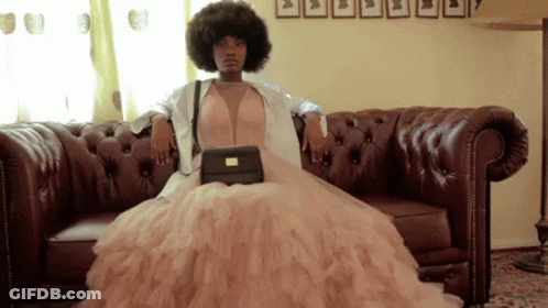 A Woman In A Dress Is Sitting On A Couch Holding A Black Purse GIF