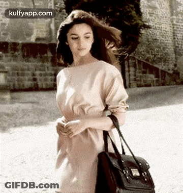 A Woman In A Dress Is Walking Down A Street Holding A Black Bag . GIF