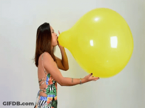 A Woman In A Floral Dress Is Blowing Up A Large Yellow Balloon . GIF