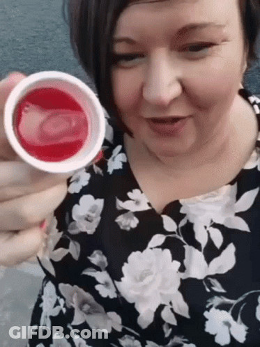 A Woman In A Floral Dress Is Holding A Cup Of Liquid In Her Hand . GIF