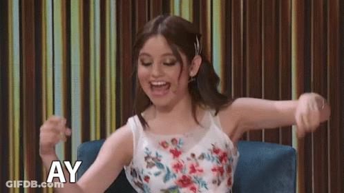A Woman In A Floral Dress Is Sitting In A Chair With Her Arms Outstretched And The Word Ay Above Her GIF