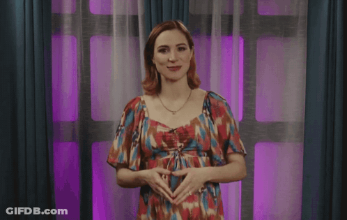 A Woman In A Floral Dress Is Standing In Front Of A Window With Purple Curtains GIF