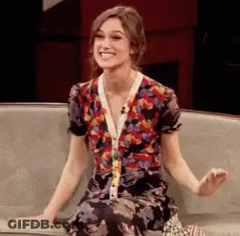A Woman In A Floral Dress Sits On A Couch Smiling GIF