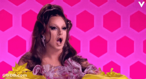 A Woman In A Floral Dress Stands In Front Of A Pink Background With A Letter V On It GIF