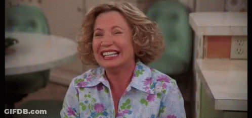 A Woman In A Floral Shirt Is Laughing In A Kitchen While Sitting At A Table . GIF